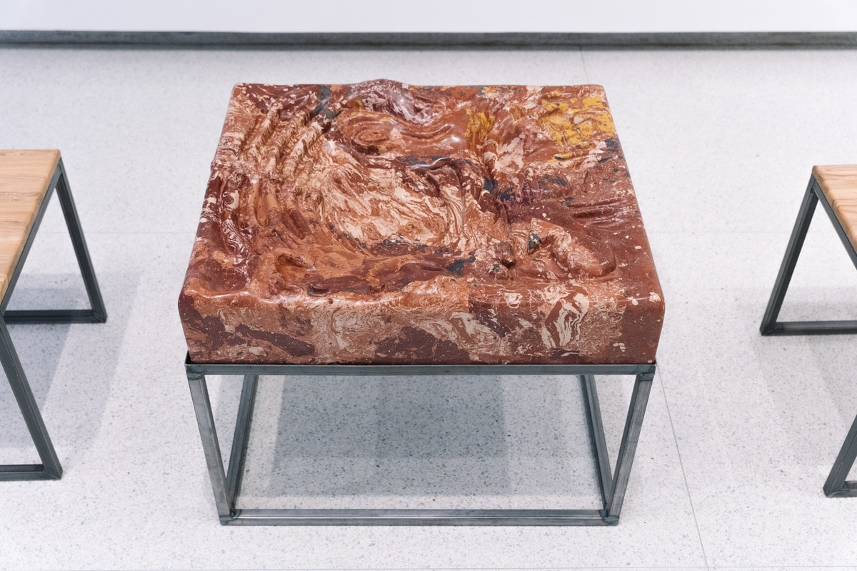  This is a photo of a “tactile theater, ” modeled after the conceptual artist Jan Svankmajer.  Svankmajer focused his artistic practice on revealing the power and evocations reveled from touch or tactile connections. The artist carved dips, divets, and grooves of various widths and depths into a plaster block for patrons to touch. The plaster has a marble pattern made of deep sienna, browns, and creams.    This tactile theater is mounted on a cube made of black metal bars. Two birch stools with legs made of the same black bars, are positioned on either side of the theater. 