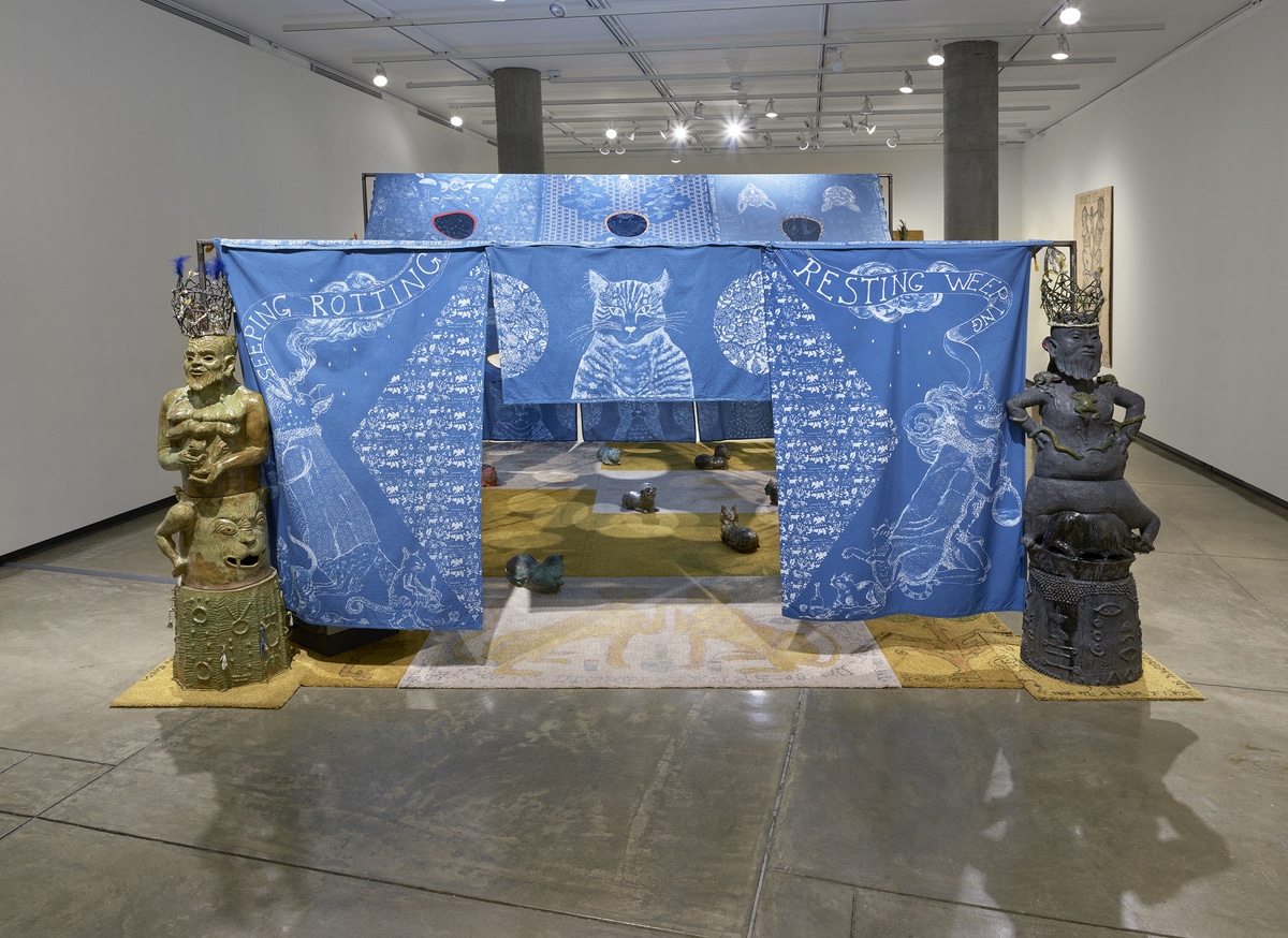 A view of the multi-part installation made of indigo dyed cotton panels from the front of the tent. The artist has batiked the indigo dyed cotton panels with images of cats and mirrored patterns of flora. Sculptures of stacked bodies-- multi-armed and multi-breasted fertility figures—flank the left and right sides of the tent. The floor underneath the tent is covered by white and gold carpets with unique designs of cat guardians and other mystical figures of the artist’s imagination. Ceramic cat shaped pillows sit sporadically. Visitors are encouraged to lay on these pillows or sit under the tent.