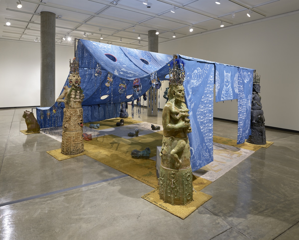 A view of the multi-part installation made of indigo dyed cotton panels from the front left corner of the gallery. The artist has batiked the indigo dyed cotton panels with images of cats and mirrored patterns of flora. Sculptures of stacked bodies-- multi-armed and multi-breasted fertility figures—flank the left and right sides of the tent. The floor underneath the tent is covered by white and gold carpets with unique designs of cat guardians and other mystical figures of the artist’s imagination. Ceramic cat shaped pillows sit sporadically. Visitors are encouraged to lay on these pillows or sit under the tent.