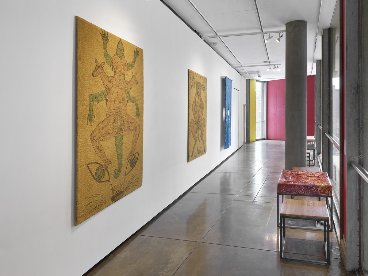 A photo of the corridor featuring three hand painted textiles hanging on the wall to the left. One of the tactile theaters, Tactile Theater #2 (after Svankmajer), 2021 is exhibited on the right. The “tactile theater,” modeled after the conceptual artist Jan Svankmajer focuses his artistic practice on revealing the power and evocations reveled from touch or tactile connections. The artist carved dips, divets, and grooves of various widths and depths into a multicolored plaster block for patrons to touch. This tactile theater is mounted on an armature made of black metal bars. Two wood-topped stools with legs made of the same black bars, are positioned on either side of the theater.